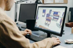 Personne de dos assise à un bureau, en train de modéliser du mobilier en 3D sur un logiciel affiché sur un grand écran